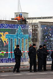 Frontale Farbfotografie von der Entfernung der Berliner Mauer mit schwarz gekleideten Polizisten und einem Absperrband im Vordergrund. In der Mitte ist ein Mauerstück nach hinten versetzt worden und ein Bauarbeiter befestigt eine Hebevorrichtung mit Ketten an dem linken Mauerstück an.