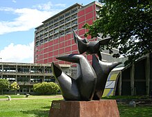 Modern outdoor sculpture, with a red building and elevated walkway in the background