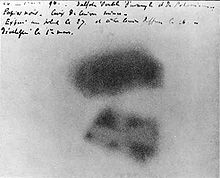Zwei verschwommene schwarze Merkmale auf einem verschwommenen weißen papierähnlichen Hintergrund.Oben im Bild befindet sich eine Handschrift.
