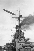 The gun turrets of a battleship. A gray zeppelin flies overhead