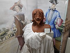 Photographie d'une vitrine. À l'arrière-plan, un fond illustré montre un homme en costume beige de mousquetaire du XVIIe siècle avec bandoulière, fraise en dentelles et chapeau à plume, et un autre en costume du XVIIIe siècle, bleu à manches violettes, portant une perruque blanche et un tricorne noir. Au premier plan, un mannequin en bois sculpté représentant un homme à moustache et barbiche fine à la façon du XVIIe siècle expose une large chemise blanche au col orné de dentelles.