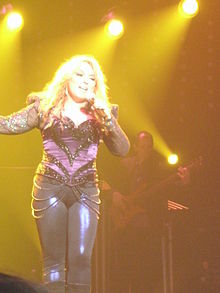 A blone woman is wearing a purple dress and black pants and is performing on a stage
