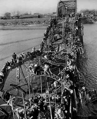 Schwarz-Weiß-Foto von Menschen, die einen Fluss über eine zerstörte Brücke überqueren