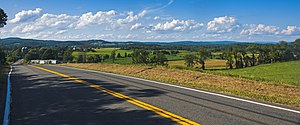 A rural landscape with a mix of fields and woodlots and low wooded mountains extends in the distance beneath a blue sky full of clouds. From bottom right to center left a paved road with a yellow stripe in the middle fills the image.