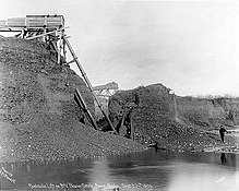 Creek-mining with hydraulic lift, 1905, showing a man beside a 50 feet high machine which has sucked up gravel from the bottom of a creek and created a brink