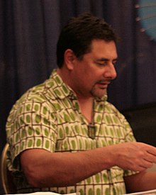 Three quarters head and waist of Lani Tupu wearing a patterned short sleeved shirt. He is doing something with his hands in front of his body and looking at them.