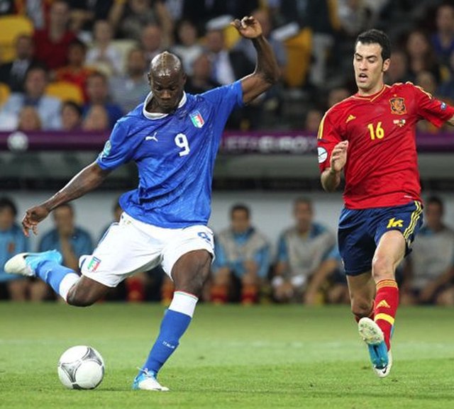 Spain holding midfielder Sergio Busquets (16, red) moves to block a shot from Italian striker Mario Balotelli.