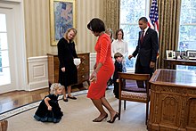 Michelle Obama and a girl curtsying - one leg ahead of the other with both legs bent. The heel is up for the back leg. The back leg crosses the front leg. Michelle Obama curtsies with Lynne Silosky.jpg