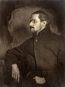 Half-length portrait of man in his thirties. He looks to his right so that his face is in profile. He has a mustache, a thin beard, and medium-length hair slicked back, and wears a pince-nez and a plain dark greatcoat, looking vaguely like a Russian revolutionary.