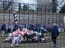 Photograph of a football scrum on a long narrow pitch with ropes and nets along the sides