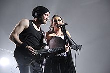 Photo en couleur montrant un homme barbu portant un bonnet jouant de la guitare et, à ses côtés, une femme brune chantant dans un micro