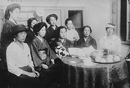 Photo de neuf femmes assises autour d'une petite table ronde. Certaines d'entre elles portent des vêtements traditionnels japonais, d'autres, des vêtements à la mode occidentale.