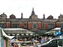 Burton Market Hall on market day. Built in 1883 to replace an older structure, the footprint of which is marked by four L-shaped metal pieces set into the ground roughly halfway between the church and the high street. The hall has a trussed roof with cast iron support pillars. Architects: Dixon & Moxon of Barnsley