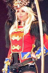 Kırmızı bir ceket giymiş olan, omuzlarında siyah tüyler ve kafasında leopar kafası şeklinde bir başlık olan sarı saçlı kadın.
