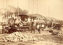 Photo ancienne d'une locomotive à l'arrêt, dans laquelle se trouve un groupe de personnes, dont de nombreux hommes coiffés de casques coloniaux.