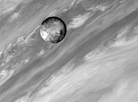 Spacecraft gray-scale photo of a planetary body (Io) covered in numerous dark spots in front of the bright and dark clouds of Jupiter.