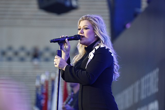 A young woman with long blonde hair wearing a dark coat and singing into a microphone