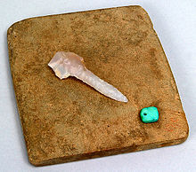 A small, translucent quartz-like chisel tool sits on a square brown tile. A small square of turquoise stone sits to its lower right.