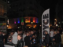 Photographie du quartier du Marais de nuit montrant une foule et l'éclairage d'un bâtiment aux couleurs du drapeau LGBT