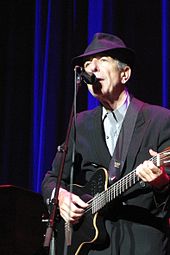 A man wearing a black fedora and a black suit. He is playing the guitar and singing into a microphone.