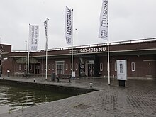 Eingangsbereich des Museum Rotterdam ’40 – ’50 NOW am Hafenbecken, mit mehreren Fahnen und einer Sitzbank vor dem Eingang