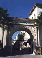 Photographie de l'arche stylisée qui marque l'entrée des studios Paramount, sous un ciel bleu, une grille fermée empêche de pénétrer dans l'enceinte, deux palmiers jouxtent l'arche.