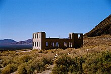 A roofless two-story masonry building rests in a setting of low shrubs and gravel under a cloudless blue sky. The building has many window openings but no glass. A mountain or hill is nearby, and a separate mountain range is visible in the distance.