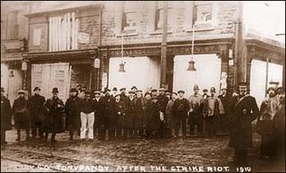  A sepia-toned photograph shows a group of men standing in front of a row of buildings with signs indicating shops or businesses. The men are dressed in early 20th-century attire, including hats and overcoats. The image is labeled "Tonypandy after the strike riot 1910”. The men appear somber, and the buildings show signs of disrepair or damage.