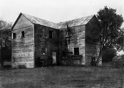 Ein Mann und eine Frau stehen vor einem alten Blockhaus. Foto aufgenommen von Albert Percy Godber.