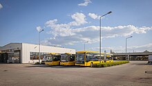 Foto von drei gelben Bussen auf einem größeren Parkplatz vor einem niedrigen grauen Gebäude