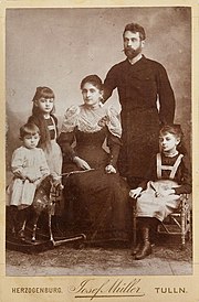 Photo sépia montrant un homme barbu debout, une femme assise en robe longue, une fillette assise, une autre debout, et un garçonnet sur un cheval de bois