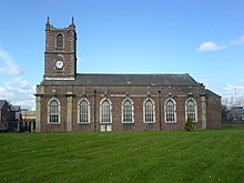 A brick church with stone dressings seen from the south. The west tower has a clock and pinnacles, and along the south face of the body of the church are Georgian-style windows