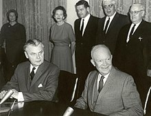 Diefenbaker i uśmiechnięty łysy mężczyzna w garniturze siedzą przy stole.  Za nimi stoją dwie kobiety i dwóch mężczyzn.