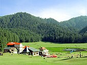 Daytime sunlit view of a verdant bowl-shaped meadow with a small pond in a valley rimmed by several prominent hills covered in evergreen forest. At centre-left and looking out on the meadow, a small cluster of tourist lodges with pitched roofs, one a bright red. Around them and the pond are small, gaily coloured gazebos and shade-giving umbrellas. Several dozen tourists and sheep mill about the pond and in the meadow.