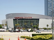A white round building that has one glass front wall showing with a sign in red text that reads"Staples Center" in capital letters. In the background, there are multiple people walking in front of the building and a white parked car and a cloudy blue sky.
