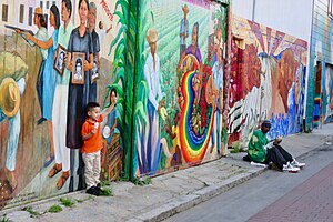 A child poses for a photo in front of a mural in Balmy Alley. Mission Mural - Political Art - 39176788725.jpg