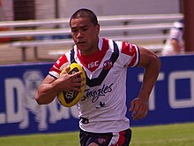 Photographie d'un homme portant un maillot de rugby blanc, bleu et rouge, tenant un ballon de rugby.