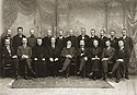 A black-and-white photograph of twenty men wearing formal clothing, nine of whom are sitting in a row on chairs while the other eleven stand behind them