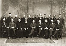 Fotografía en blanco y negro de 20 hombres posando con traje ceremonial en una habitación ricamente decorada.