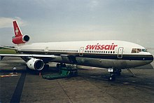 Foto ingrandita di tre quarti della parte anteriore di un aereo di linea visto sul lato destro.  L'aereo, nei colori bianco e rosso con una croce bianca su sfondo rosso sulla coda, è parcheggiato sull'asfalto di un aeroporto, con il portello di carico aperto.