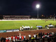 Un meci de rugby jucat noaptea, pe un stadion luminat.  Spectatorii din tribună și din jurul terenului sunt toți îmbrăcați în alb și roșu.