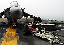 Dos miembros de la tripulación inspeccionando una bomba en un carro, frente a un avión.