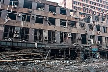 Photographie couleur. Bâtiment haut de trois étages à la toiture partiellement détruite, aux vitres brisés et à la façade noircie, au milieu d’un paysage urbain sinistré. Présence d'un branchage d'arbre couché et de nombreux débris au sol au premier plan