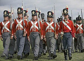 Foto di uomini vestiti con uniformi britanniche dei primi del 1800.  Indossano cappotti rossi con cinture incrociate bianche, pantaloni grigi e shakos neri.