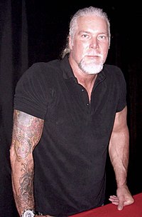 Homme blanc aux cheveux gris et tatouages ​​aux bras portant une chemise noire