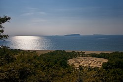 Suuri kolikkoa esittävä hiekkaveistos Seton sisämeren rannikolla, taustalla Ibukijima