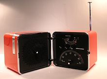 Radio.  Red plastic cube with an edge length of about 15 centimeters, unfolded.  The black cut surfaces are visible and show a loudspeaker on the left and the radio controls (buttons and dials) on the right.  On the right a half-extended telescopic antenna.