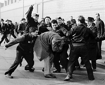 alt = A black-and-white photograph of a group of 4–6 members of United Auto Workers beating a man. The man is trying to defend himself by covering his head with his coat.