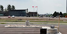 A building on a clear day. It is situated amongst considerable asphalt, with trees in the bacground. Two flag poles are in front of the building, an Ontario and Canada flag. The building is designed with particular attention to obscure angles.