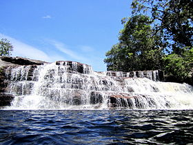 Photograph of a wide, blue pool underneath a cascading, stepped waterfall fringed by mature, tropical trees